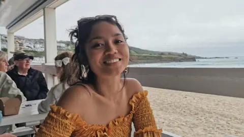 A woman in a yellow off-the-shoulder top smiles at the camera while sitting at a table on a beach overlooking the ocean. 