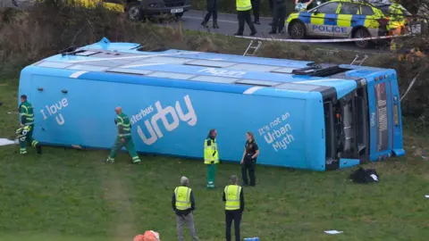 PA A blue Ulsterbus lies on its side in a field. There are emergency workers standing around and some discarded clothing. A police car and an ambulance are on the road. Police, fire fighters and ambulance service workers are there.