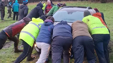 A group of eight people wearing waterproofs pushing a car in a muddy field.