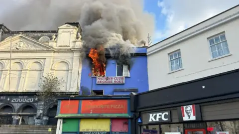 A blue multi-storey building is seen with flames coming from its top floor. There is a sign reading National Club Reggae Soul beneath the blazing windows. To the left of the building is a KFC. 