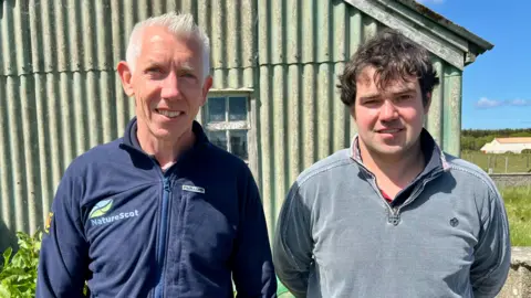 Robbie Kernahan stands wearing a NatureScot top beside farmer Douglas Paterson, outside in bright sunshine against a light green corrugated iron shed.