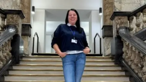 Agata Wiznefska Agata Wiznefska is smiling at the camera. She has shoulder length dar hair and is wearing a dark jumper over a white shirt and blue jeans. She is standing on a set of marble steps.