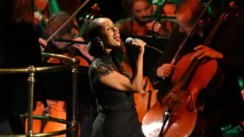 A woman, Andrea Lykke, in a black dress sings into a mic. Cellists from an orchestra are behind her