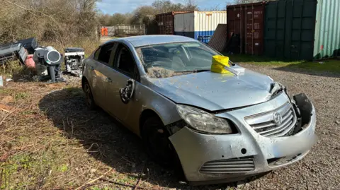 A car with a damaged bumper is abandoned in a grassy site by a train station. It is silvery-grey, and has dust and dirt on its front window. There is yellow equipment on the bonnet. Behind are large shipping containers.