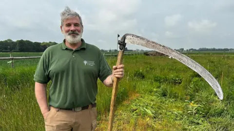 Andrew Callaghan, who has grey hair and a grey beard, standing in a field holding a scythe in his left hand. He is wearing a green short-sleeved shirt with a "Fenland Orchards Project" logo and wearing light brown trousers.