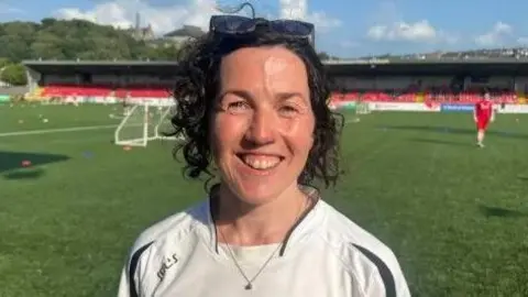 Meabh O'Neill stands in the middle of a football pitch. An empty stand filled with red seats is in the background. A lone figure dressed in a red kit walks towards her. The woman is standing in front of a set of goals and is wearing a white football shirt with the words Foyle pride written on it.
