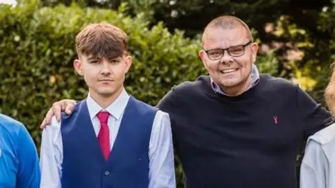 Martin Cosser Close up image of Charlie Cosser and Martin Cosser. Charlie is wearing a tie and waistcoat, while Martin is wearing a dark-coloured jumper. They are stood outside and smiling at the camera.
