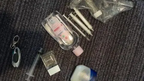 CPS Syringes, a tub of Vaseline and a bottle of baby oil on a dark grey carpet.