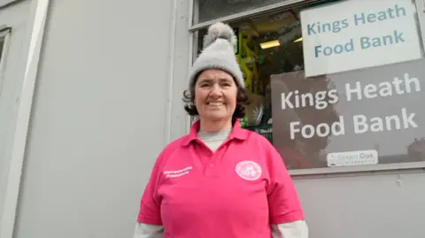 Kings Heath Food Bank coordinator Sharon Power stands outside the storage unit for the charity wearing a pink polo shirt with the food bank's insignia and a sponsor's logo for Worcestershire Freemasons on top of a charcoal grey jumper. On top of her short brown hair, she is wearing a tall, grey bobble hat