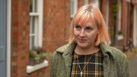 Sam Read/BBC Jennifer Wilson-Marklew with medium-length orange hair looking to the left of the camera and wearing a green coat over a green and brown top. She has a small pendant on a necklace around her neck and is standing in front of brick-built houses with planters on the exterior ledges.