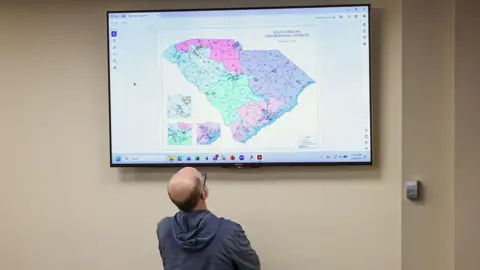 Bloomberg via Getty Images A man, seen from behind and wearing a gray hooded sweater and glasses, looks up at a proposed Congressional map for South Carolina displayed on a screen on a beige wall. 