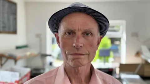A man pictured from the shoulders up, wearing a hat and a pink shirt pictured inside a room, with a blurred background behind him