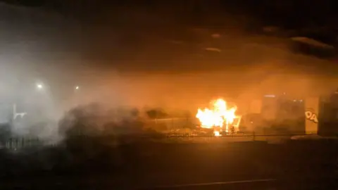 BBC Pictures of the campervan fire. The vehicle is completed engulfed in orange flames. Thick smoke has gathered under the bridge.