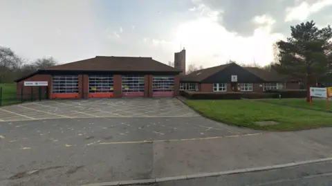 Google A single-storey fire station building with four large doors for engine access and tree to the right 