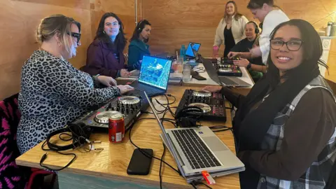 Cad Taylor/BBC A table of women working on DJ equipment. Two women are looking towards the camera