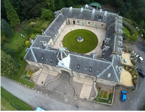 Falkland Estate Aerial view of a large stone house with a square layout surrounding a circular lawn courtyard, set among trees and gardens.
