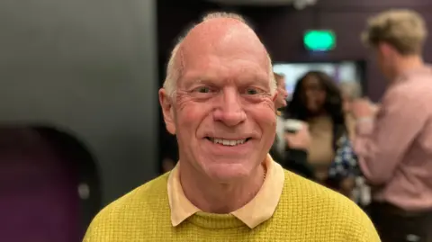 Laura Coffey/BBC Stephen Giles-Medhurst wearing a yellow textured jumper and a light-coloured collared shirt is pictured inside a venue with dark walls and bright lighting. Several people are visible in the blurred background, standing and talking near what appears to be a reception or event area.