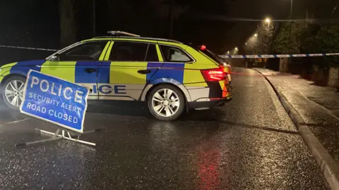 A PSNI car parked across a road with a sign in front of it saying 'Police Security Alert Road Closed'