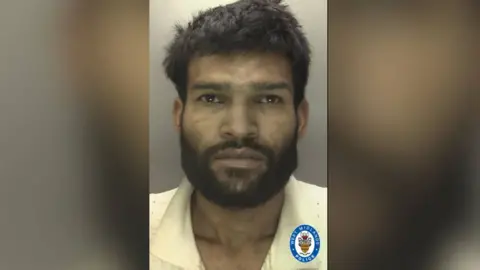 Keren Kumar is pictured in a mugshot from West Midlands Police with no expression on his face. He has messy black hair and a full black beard.
