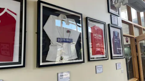 Various signed sports tops and t-shirts in frames on a white wall. Below them are names with information. There is a window and door to the right of the image.