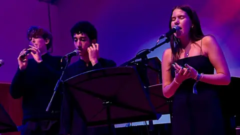 Cad Taylor /BBC Three musicians stand in front of three microphones one on recorder and two singing
