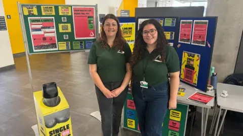 George King Two women wearing dark green polo shirts standing in front of boards on which there are posters about a charity's Single Use Sucks campaign against disposable vapes