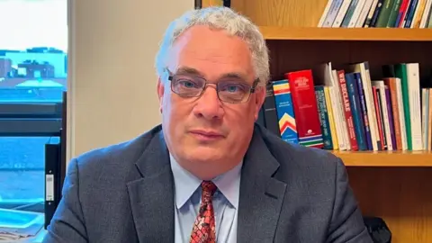 Aengus Ó Snodaigh A man with grey hair and glasses is wearing a blue jacket over a blue shirt and red tie. There are bookshelves behind him with assorted hardback and paperback books.