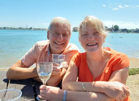 Family photo Chris and Ruth Stone-Houghton sit on a picnic bench, smiling into the camera. They are holding alcoholic drinks, and behind them is a body of water