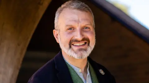 Headmaster of Bedales, Will Goldsmith, in front of the school door. He is smiling and has short grey hair and a grey beard. He is wearing a navy jacket over a blue and green shirt. There is a white badge on his lapel. 