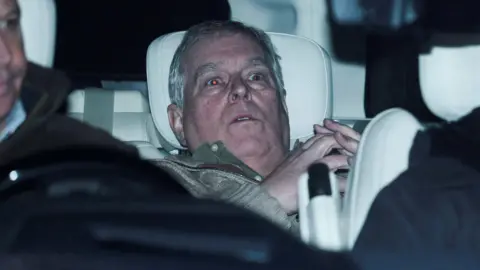 A horizontal grey-haired man looking alarmed in the back seat of a car
