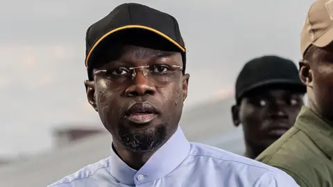 A head and shoulders shot of Ousmane Sonko wearing glasses and in a black cap and buttoned-up blue shirt.