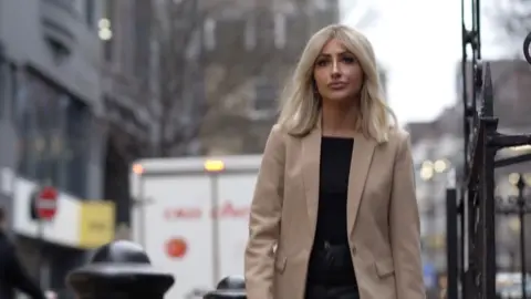 Holly is walking in the street, with a beige blazer and black top and jeans. She has blonde hair and is looking past the camera. 