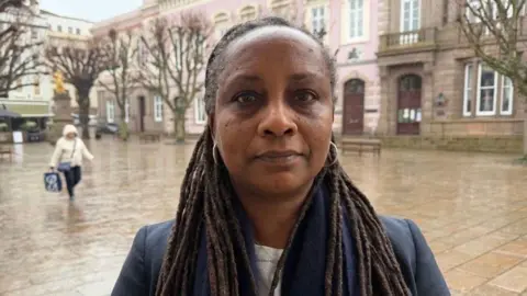 BBC Deputy Beatriz Porée is pictured in Jersey's Royal Square. She is wearing a blue scarf and a black blazer. 