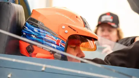 Matthew Willis Close-up of Dave Warby in the Bluebird cockpit. He is wearing an orange crash helmet, which is decorated with an image of the Australian flag and his own name.