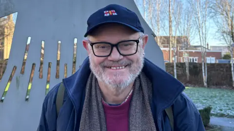 Sophie Montague/BBC Brian Kell is smiling directly at the camera while standing in front of a metal 2D Straw Bear sculpture. He has black square framed glasses and grey facial hair. He wears a blue cap with the RNLI's lifeboats logo in the centre. He also wears a blue coat and red jumper - both branded with a Straw Bear.