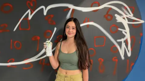Rebecca Baldwin a young girl with brown hair stood in front of a creatively painted wall and holding a white spray paint cannister
