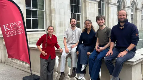 King's College London A picture of the research team - three men and two women - outside a stone university building. Four of the team sit on a wall, while Dr Crane wearing a red jacket stands next to a flag with the words King's College London on it.