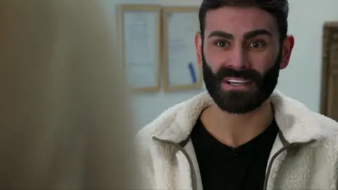 Matthew Wood, who has brown hair and a brown beard, speaks to the camera wearing a white woolen fleece