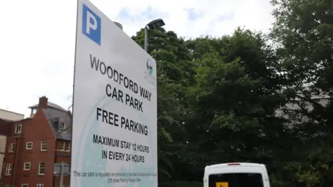 A close-up shot of a sign that reads: 'Woodford Way car park. Free parking. Maximum stay 12 hours in every 24 hours.'