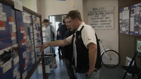 Shaun Whitmore/BBC Actor Josh Bolt is wearing a white shirt and black sleeveless jacket and standing inside the museum, pointing at an artefact on the wall.