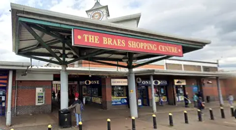 Google The entrance to The Braes Shopping Centre features a large red and gold sign beneath a wide, metal‑framed canopy. People walk in and out of the brick‑fronted shops beneath the sheltered walkway. The One-O-One off licence sits on the corner.