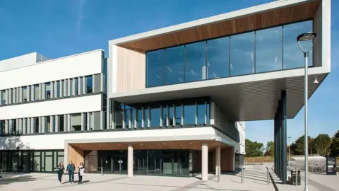 A modern university campus with three storeys and a very large top floor which projects from the rest of the building with large glass windows and wood panelling. It is a sunny day with a clear blue sky and three people are walking away from the building.