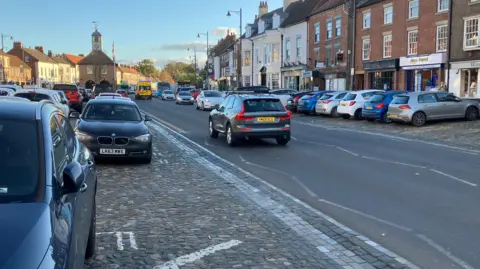 Yarm High Street. To the left there are parked cars, to the right, a row of shops and businesses.