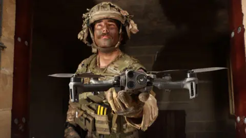 Ministry of Defence A military drone operator in camouflaged combat fatigues and helmet holds a drone with four propeller arms in his upturned, outstretched palm. He's standing inside the doorway of bare, concrete shelter.