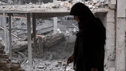 NurPhoto via Getty Images A woman in dark-coloured clothes with her head covered stands in the foreground of the picture. Behind her are grey girders that are all that remains of a building, with grey rubble and dust heaped around them. Photo: 15 March 2026.