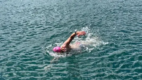 Fundraiser Jay Goss, pictured swimming front crawl stroke in the sea. He is wearing goggles and a bright pink swimming cap. 