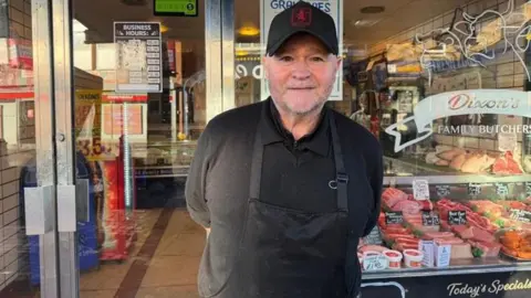 BBC Man stood in front of butchers wearing a black cap, black top and black apron