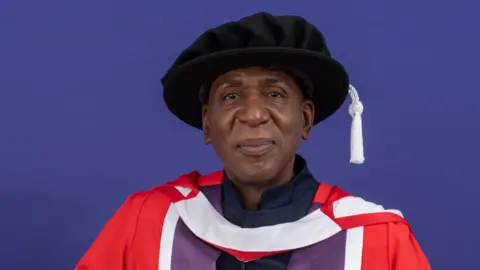 Head and shoulders of Colin McFarlane in a red and purple gown with a black hat on.