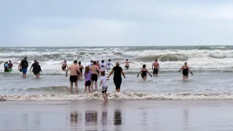 Seacoast Dippers A large group of people wading into rough sea water together during a winter beach swim.