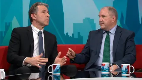 Politician Dan Norris sits on the BBC Politics West sofa as he is challenged by another guest, Simon Hoare. Both men are wearing suits and ties and there is a glass-topped coffee table in front of them with four mugs on it